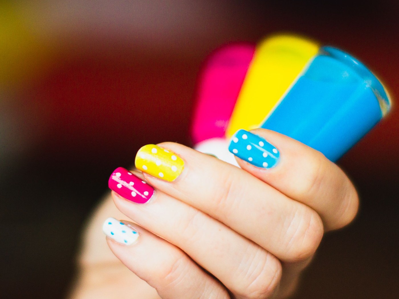Mano con uñas pintadas de rosa, amarillo y azul junto a tres esmaltes de colores
